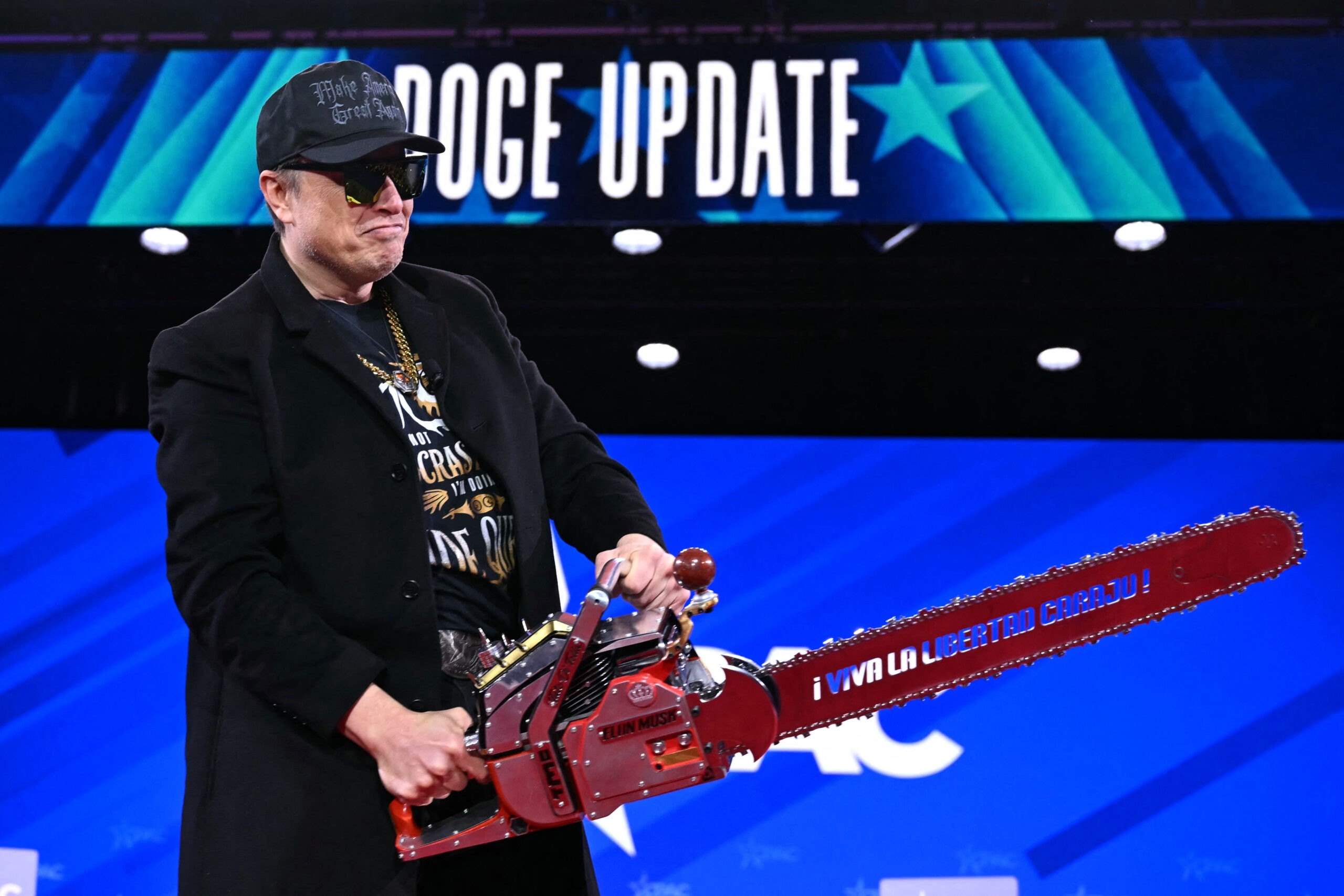 Elon Musk with a black make america great again hat, a goldchain and, a chainsaw for a doge update